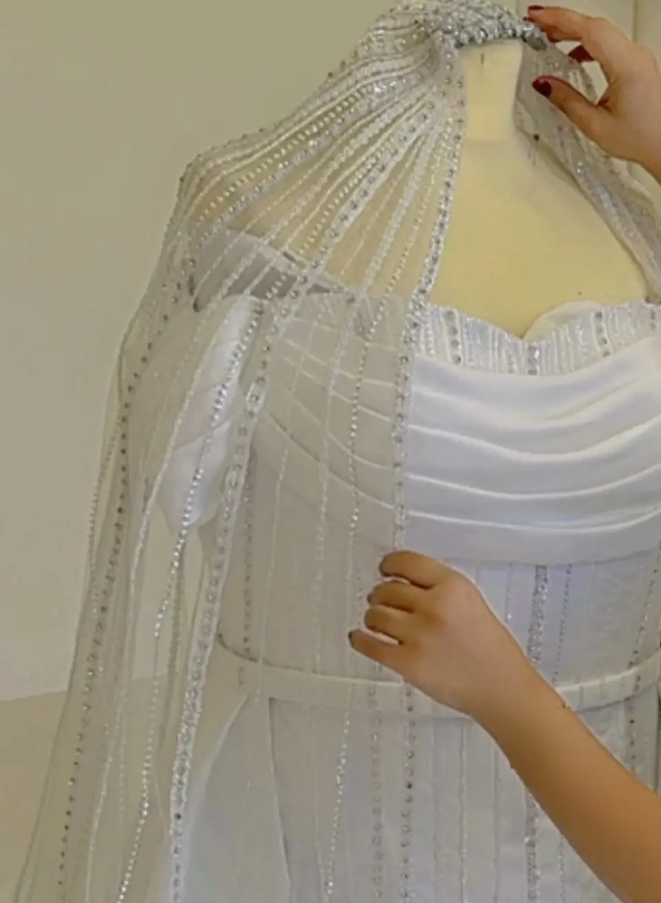 A couturier fitting a crystal-beaded tulle cape over a draped bridal bodice on a dress form