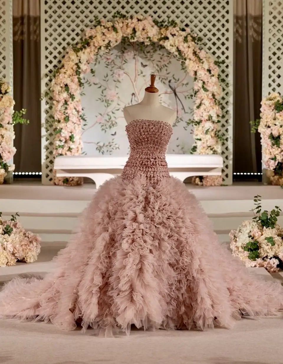 Blush couture gown with a sculpted petal bodice and cascading ruffled tulle skirt, displayed before a floral arch